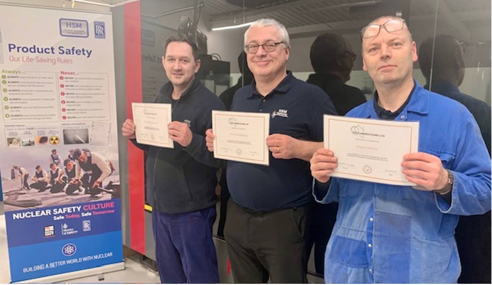 Three men holding certificates in front of a ‘Product Safety’ and ‘Nuclear Safety Culture’ poster. The poster includes safety guidelines and branding from HSM and Rolls-Royce. They are standing in a workplace setting, smiling while presenting their certificates.